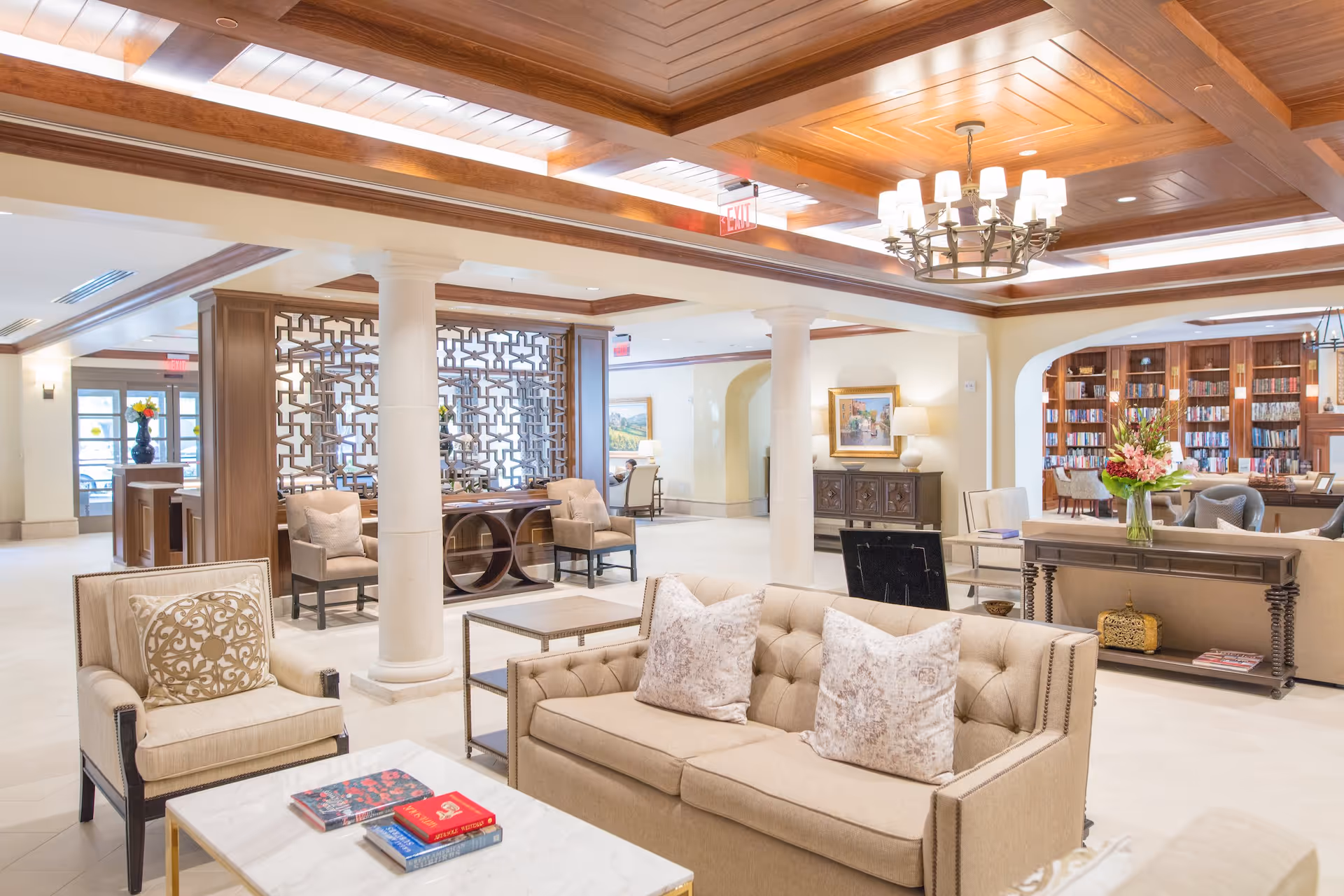 A spacious and elegant living room area with beige sofas and armchairs, a marble coffee table with books, decorative pillows, and a vase of flowers. The room features wooden ceiling beams, white columns, a chandelier, and a bookshelf filled with books in the background.