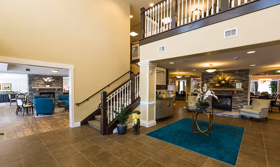 Spacious and well-lit interior of an assisted living facility featuring a tiled floor entrance area with a teal rug and a round table holding a floral arrangement. To the right, there is a cozy seating area with armchairs and a stone fireplace decorated with flowers. A staircase with dark wood railing and white balusters leads to an upper floor. In the background, additional seating areas with chairs and tables are visible, along with warm lighting fixtures.