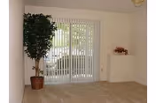 Carpeted living room with a sliding glass door covered by vertical blinds, a potted plant, and a small decorative ledge.