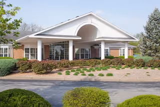 Front exterior view of a single-story brick building with white columns and a covered entrance, surrounded by neatly trimmed bushes and landscaping under a clear sky.