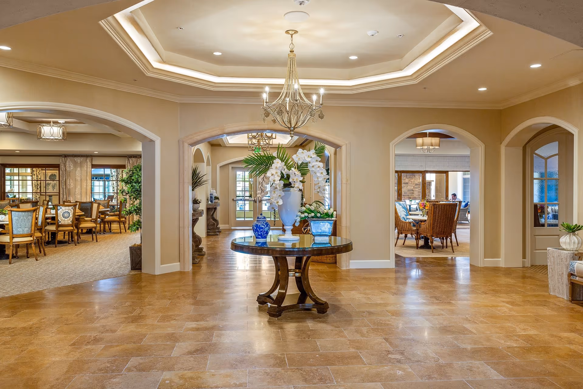 Spacious and elegant interior of The Kensington Redondo Beach featuring a central round wooden table with decorative vases and white orchids. The room has beige walls, tiled floors, recessed lighting, and a chandelier hanging from a tray ceiling. Arched doorways lead to dining areas with wooden chairs and tables, and large windows allow natural light to fill the space.