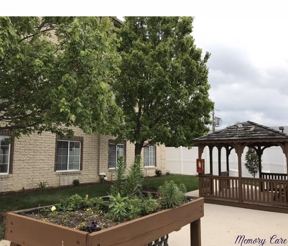 Outdoor garden area at a senior living facility with a raised planter box containing various plants, a large leafy tree, a wooden gazebo, and a beige brick building with windows in the background under a cloudy sky.
