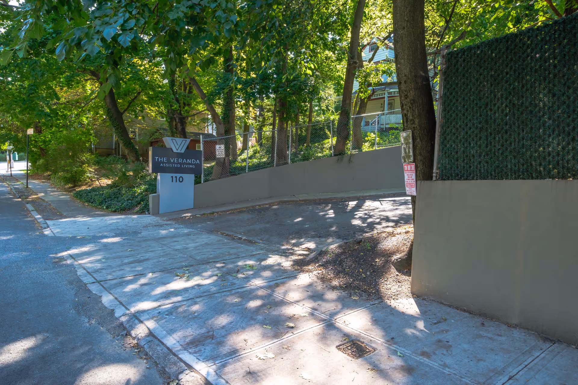 Outdoor view of the entrance to The Veranda Assisted Living facility, showing a sign with the facility's name and address number 110, surrounded by trees and greenery along a sidewalk and driveway.