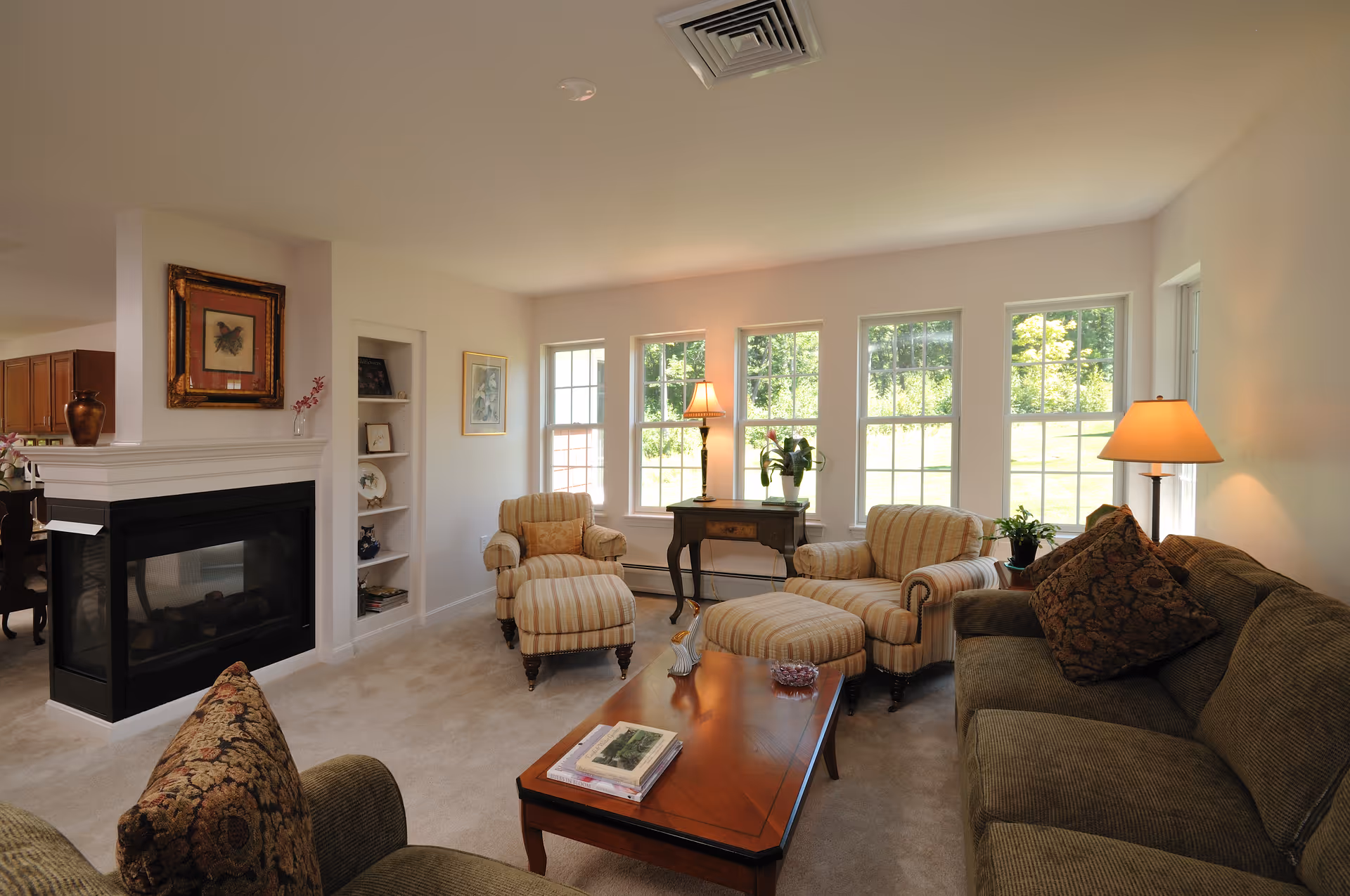 A cozy living room with a green sofa and two striped armchairs with matching ottomans. A wooden coffee table with books and decorative items sits in the center. There is a fireplace on the left wall with a framed picture above it, and a small built-in shelf next to it. Large windows on the far wall let in natural light, with a small table and two lamps placed in front of them. The room has light-colored walls and carpeted flooring.