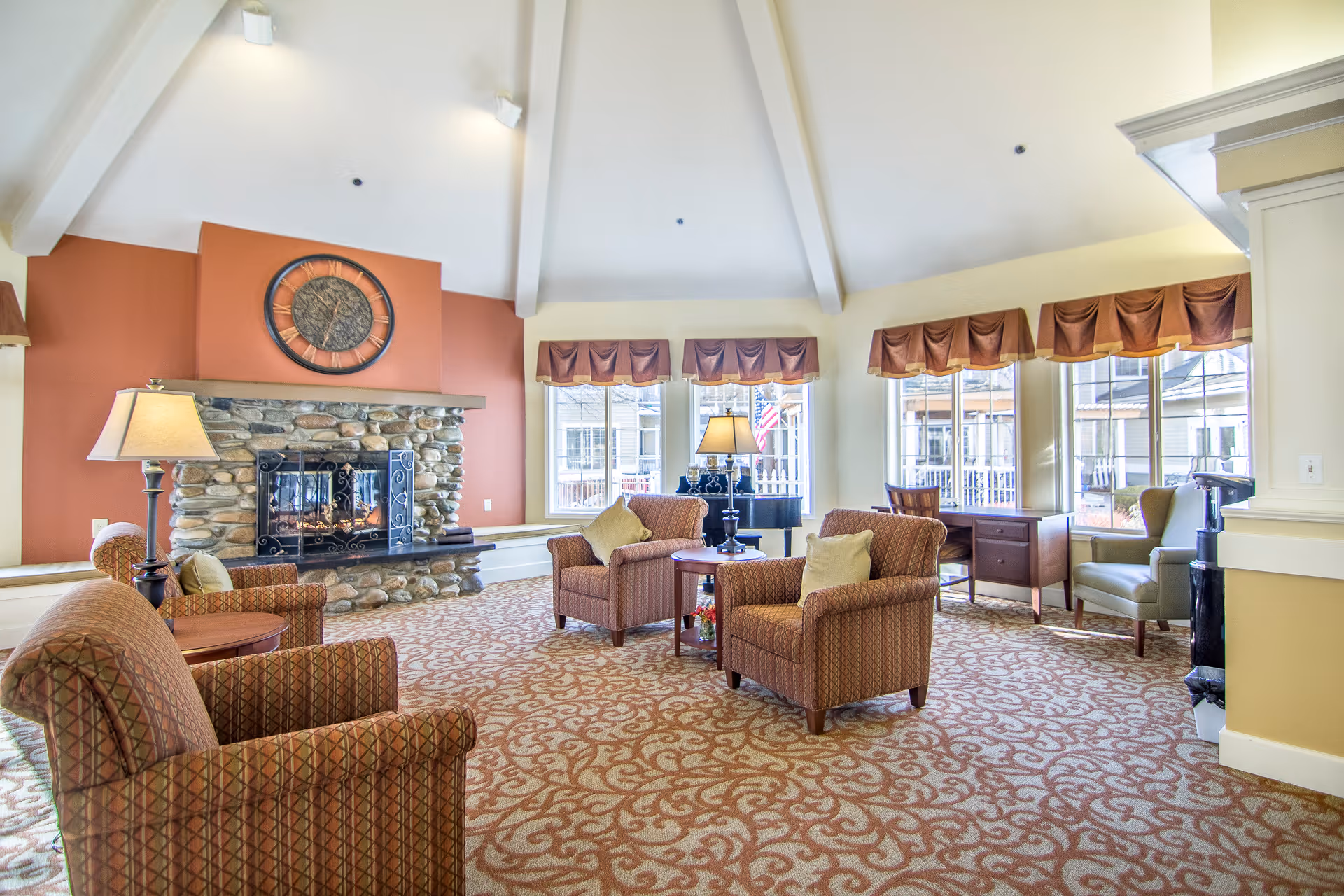 A spacious living room area with a high vaulted ceiling and large windows letting in natural light. The room features a stone fireplace with a large clock above it, several upholstered armchairs with cushions arranged around small wooden tables, a floor lamp, and a desk with a chair near the windows. The carpet has a patterned design, and the window valances are brown with a gold trim.