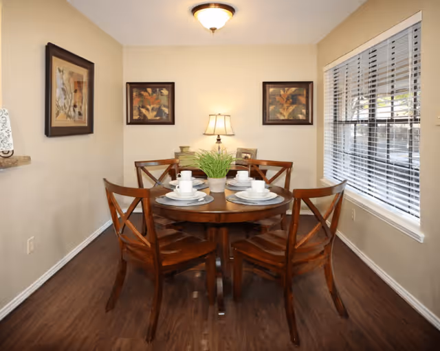 A dining room with a round wooden table set for four with white plates, cups, and saucers. Four wooden chairs surround the table. A small green plant is in the center of the table. The room has beige walls with three framed floral paintings and a table lamp on a sideboard against the back wall. A window with white blinds is on the right side, and the floor is dark wood.