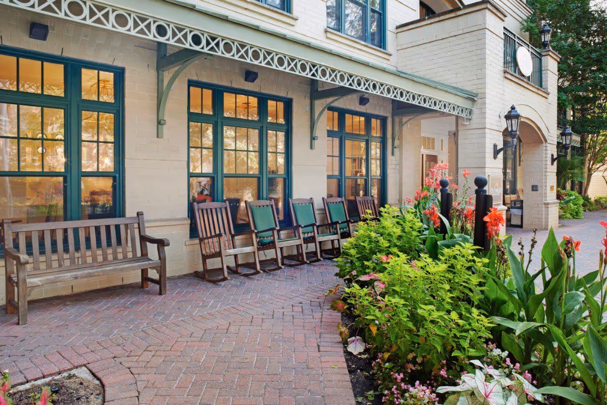 Outdoor patio area of a senior living facility with a row of wooden rocking chairs and a bench along a brick wall with large windows. There is a garden bed with green plants and colorful flowers next to the paved walkway leading to the building entrance.