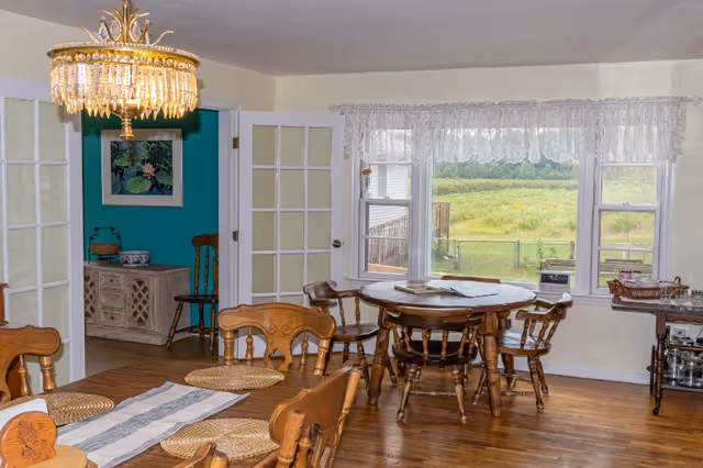 Sunlit dining room with wooden tables and chairs, a crystal chandelier, French doors, and a large window overlooking a grassy yard.