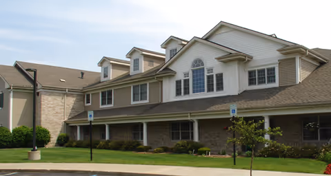 Exterior view of a multi-story senior living facility building with beige siding and brick accents, surrounded by a well-maintained lawn and small trees under a clear sky.