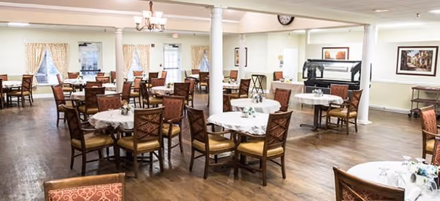 A spacious dining room with multiple round tables covered with white tablecloths, each surrounded by wooden chairs with cushioned seats. The room has wooden flooring, cream-colored walls, large windows with curtains, and decorative wall art. There are white columns and a chandelier hanging from the ceiling.
