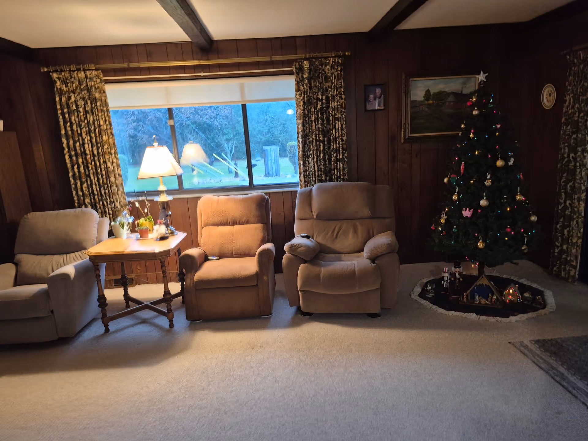 A cozy living room with three recliner chairs arranged in a row. A wooden side table with a lamp and small potted plants sits between two of the chairs. The room has wood-paneled walls and a large window with patterned curtains. A decorated Christmas tree with ornaments and lights stands in the corner on a small rug, with a nativity scene and other holiday decorations underneath. There are framed pictures and a painting on the walls.