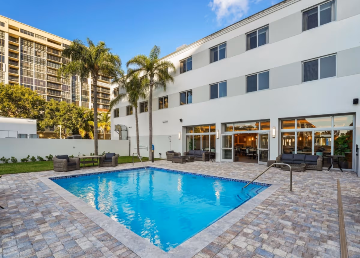 Outdoor swimming pool area at Seaside Hallandale Beach Senior Living with palm trees, patio seating with cushioned chairs and tables, and a white multi-story building in the background under a clear blue sky.