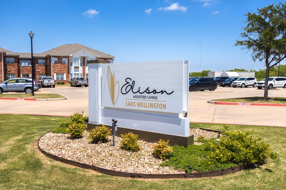 Outdoor view of the entrance sign for Elison Assisted Living of Lake Wellington, with a landscaped area featuring small bushes and rocks in front of the sign. In the background, there is a parking lot with several parked cars and a multi-story brick building under a clear blue sky.