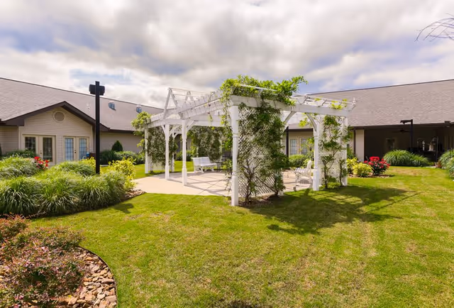 Outdoor garden area at The Bungalows at Fayetteville featuring a white pergola with climbing plants, surrounded by green grass and landscaped bushes, with a building in the background under a partly cloudy sky.