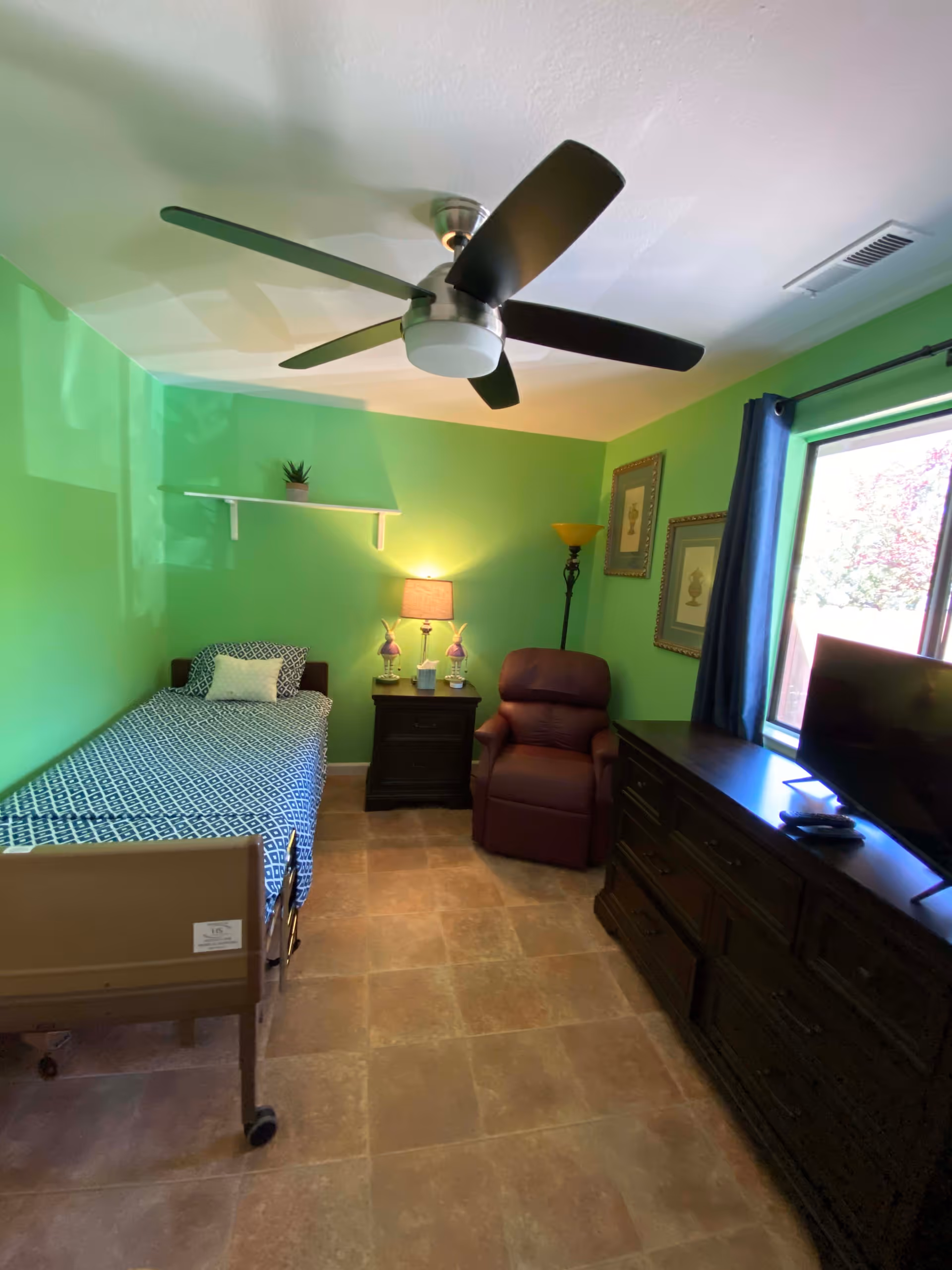 A small bedroom with bright green walls, a single bed with a patterned blue and white bedspread, a dark wooden nightstand with a lamp and decorative items, a brown recliner chair, a dark wooden dresser with a TV on top, two framed pictures on the wall, a window with blue curtains, and a ceiling fan with black and silver blades.