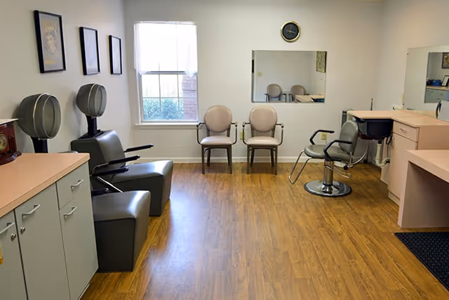 Interior view of a salon area with two hair drying chairs on the left, two beige chairs against the wall under a window, and a styling chair in front of a mirror on the right. The room has wood flooring, light-colored walls, and cabinets with a countertop.