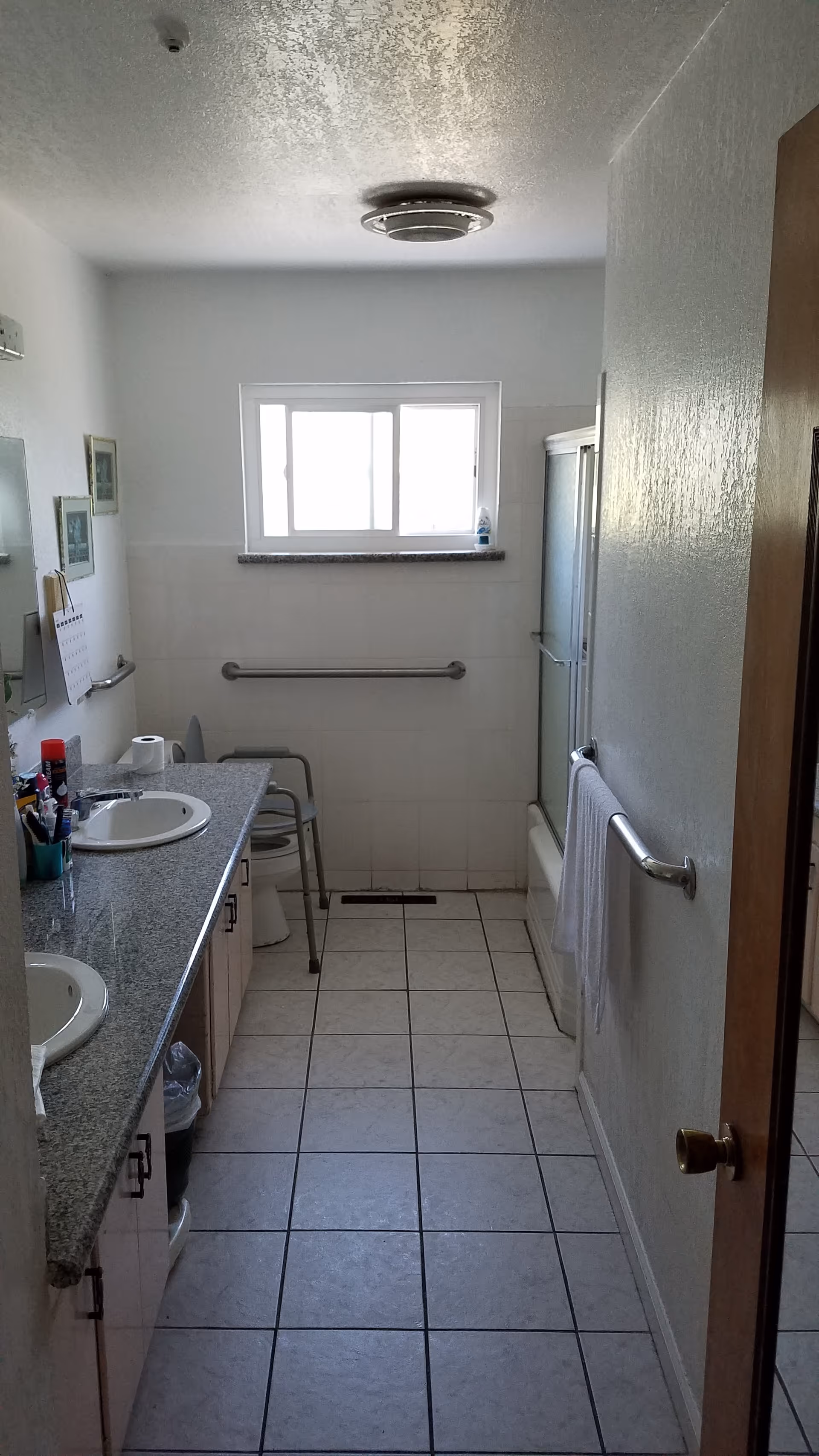 Narrow accessible bathroom with double sinks, tiled floor, grab bars, a shower/tub, and a walker near the toilet.