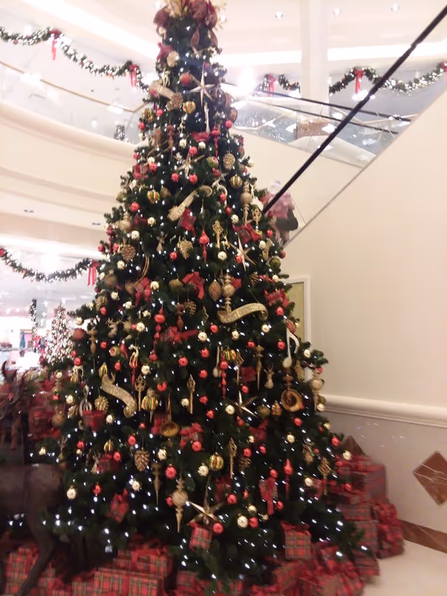 A large, decorated Christmas tree adorned with red, gold, and white ornaments, ribbons, and lights, surrounded by wrapped presents at the base. The tree is located indoors in a spacious area with a staircase and garlands decorated with red bows in the background.