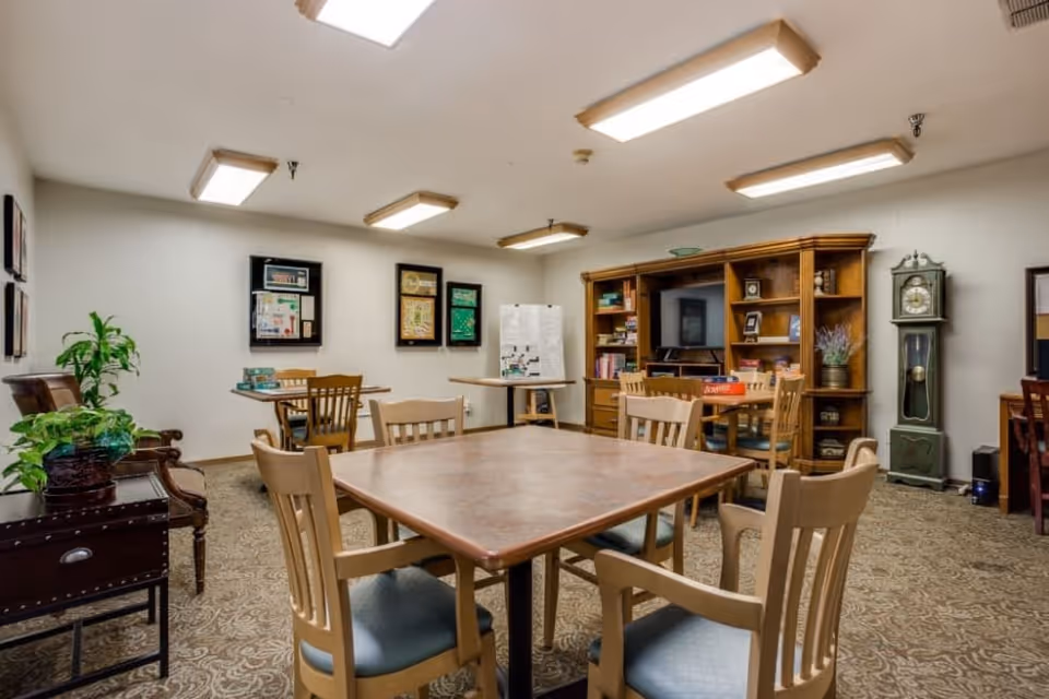 A cozy common room with a square wooden table surrounded by six chairs in the center. The room has carpeted flooring, several framed pictures on the walls, a wooden bookshelf with a TV and various items, a grandfather clock, and a few plants placed on furniture. The ceiling has multiple rectangular fluorescent lights.
