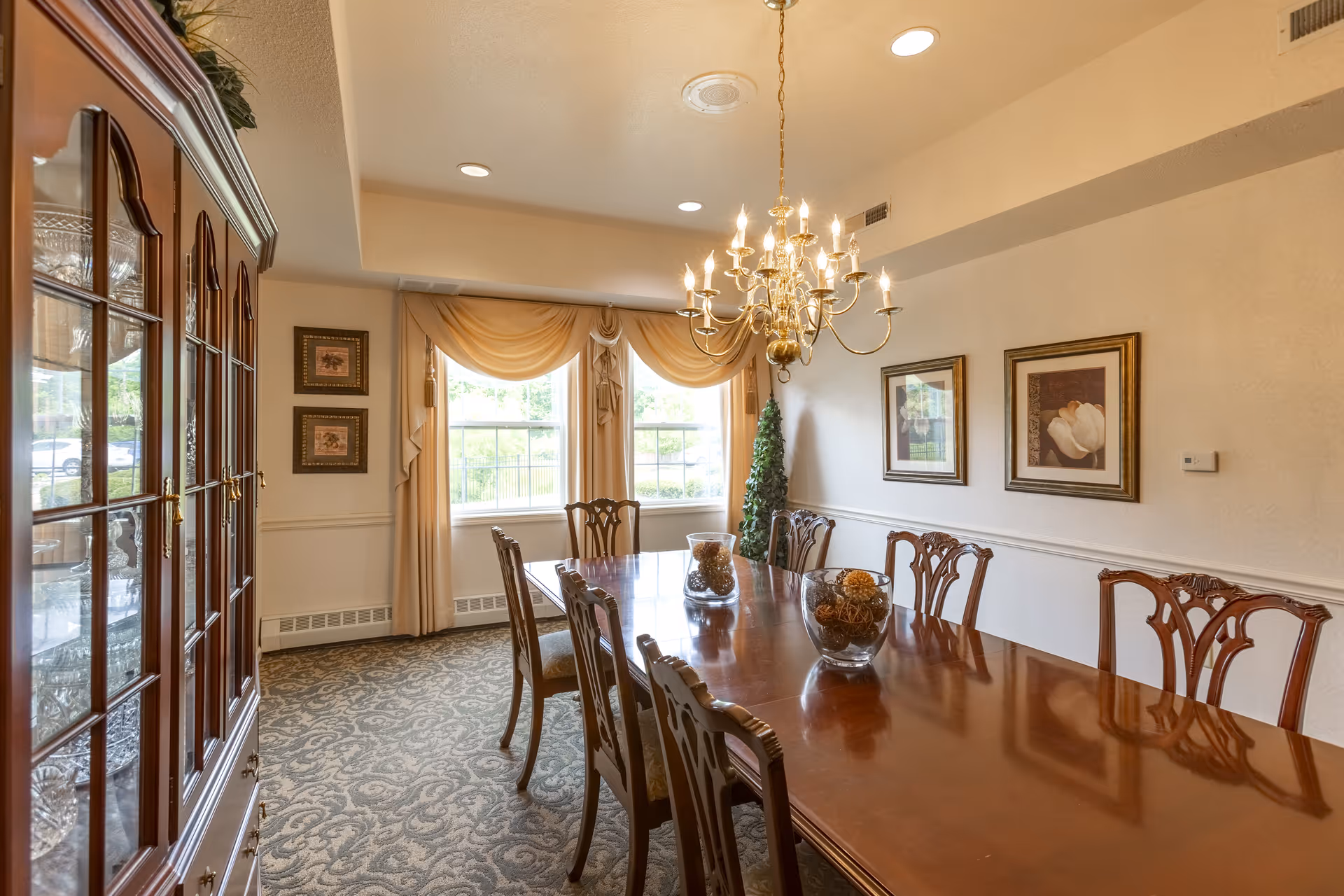 A formal dining room with a long polished wooden table surrounded by eight matching chairs. The room features a large chandelier hanging above the table, two windows with beige curtains, framed artwork on the walls, a decorative cabinet with glass doors on the left, and a small artificial tree in the corner.