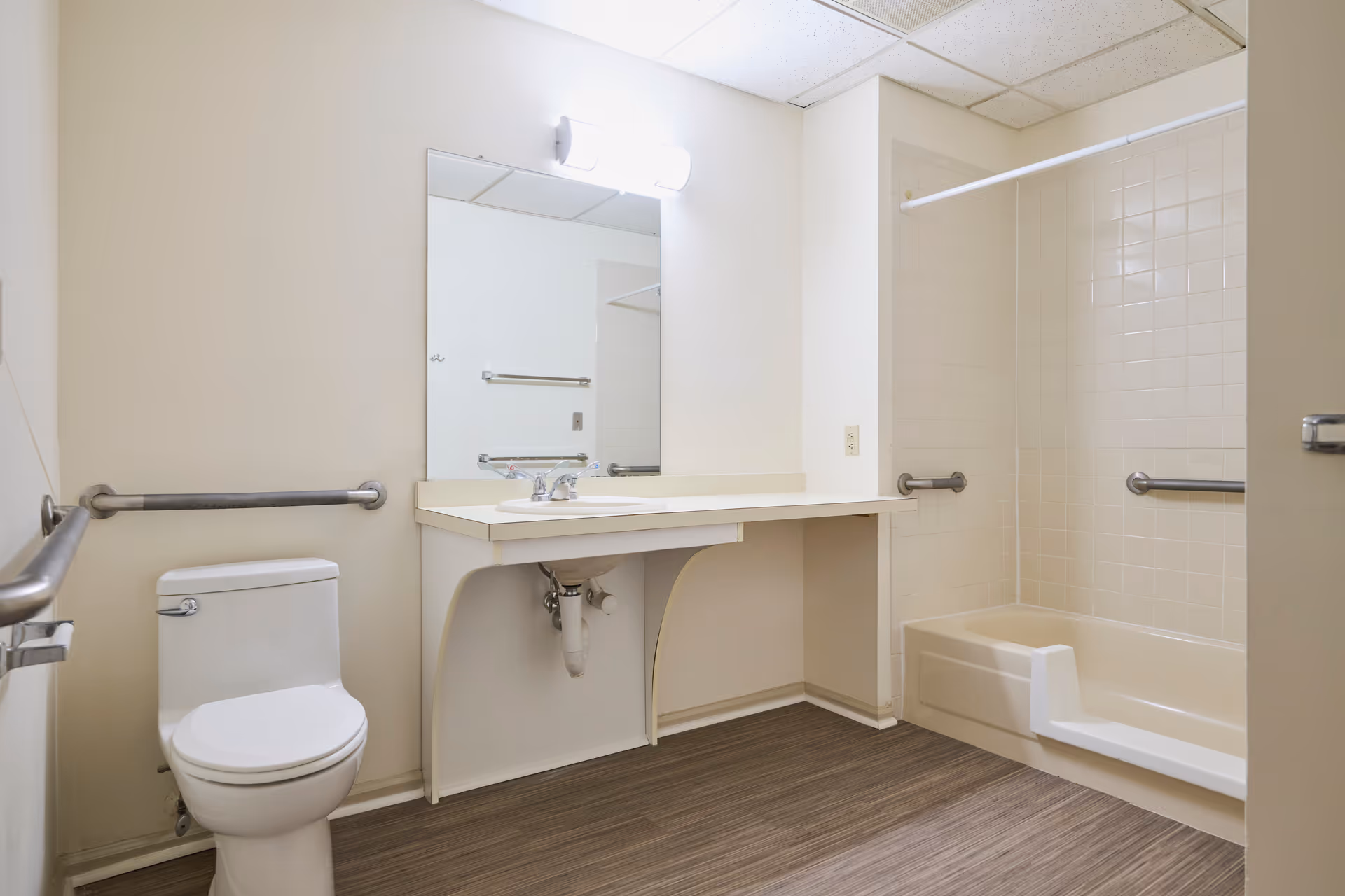 A clean and accessible bathroom featuring a toilet, a sink with a large mirror above it, and a bathtub with a shower curtain rod. The bathroom has grab bars installed near the toilet and inside the bathtub area for safety. The floor has a wood-like finish, and the walls are painted white.