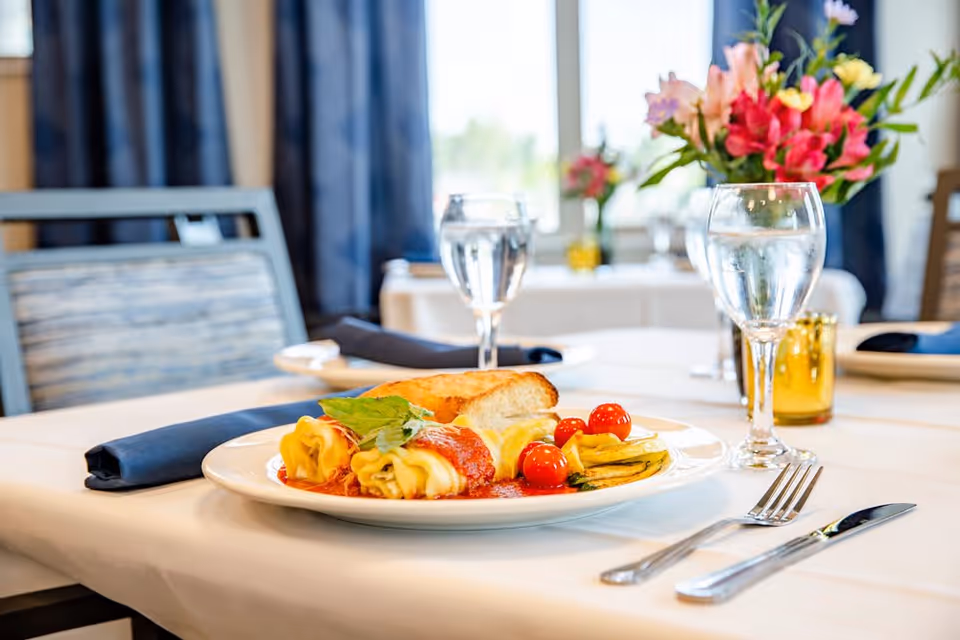 A dining table set with a plate of rolled pasta with tomato sauce, cherry tomatoes, grilled vegetables, and a piece of bread. The table is covered with a white tablecloth, and there are two glasses of water, silverware, a navy blue napkin, and a vase with pink and yellow flowers in the background.