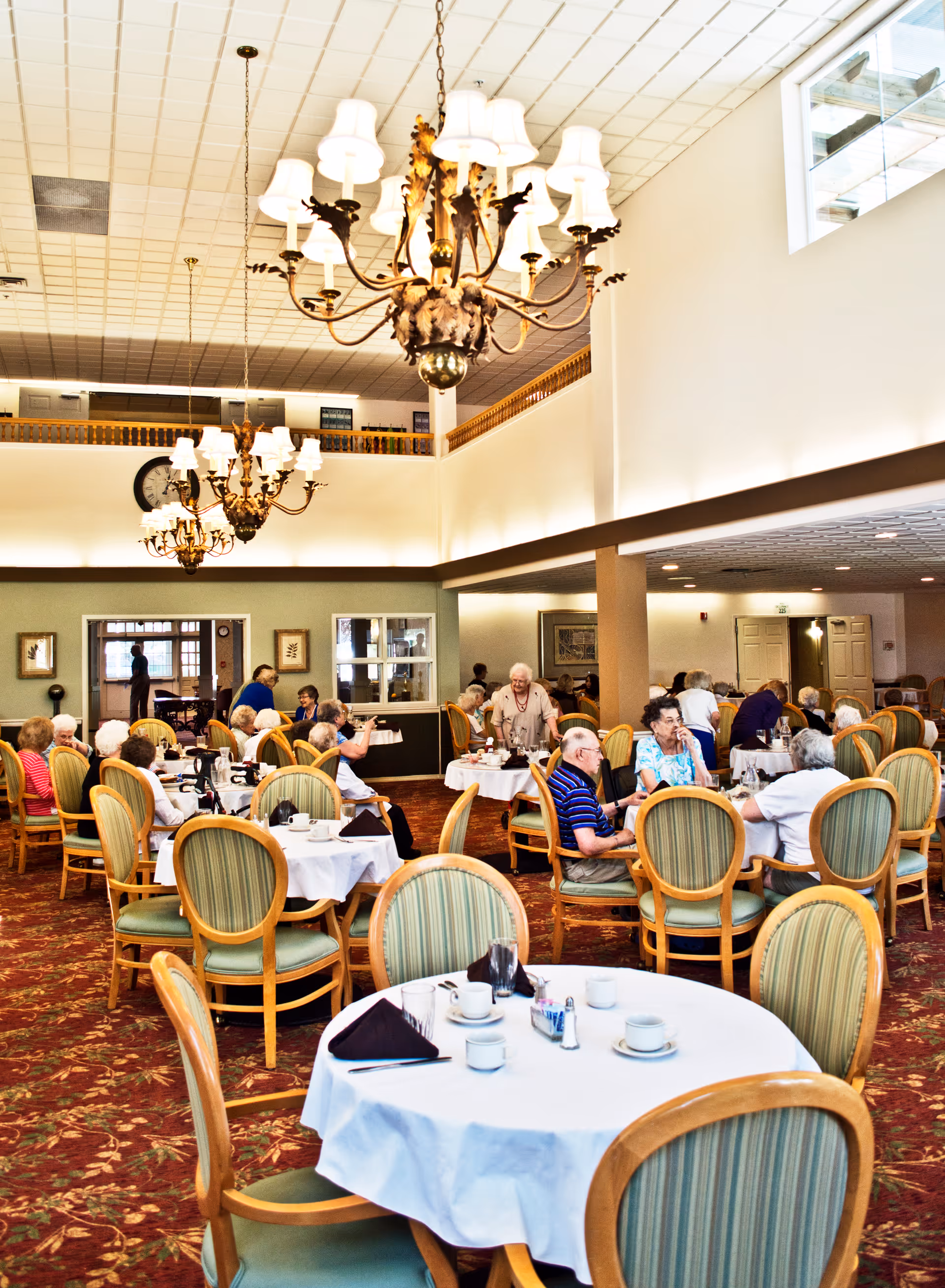 A spacious dining room in a senior living facility with multiple round tables covered with white tablecloths and set with cups, saucers, and napkins. Several elderly residents are seated and engaged in conversation. The room features high ceilings with ornate chandeliers and large windows allowing natural light to enter.