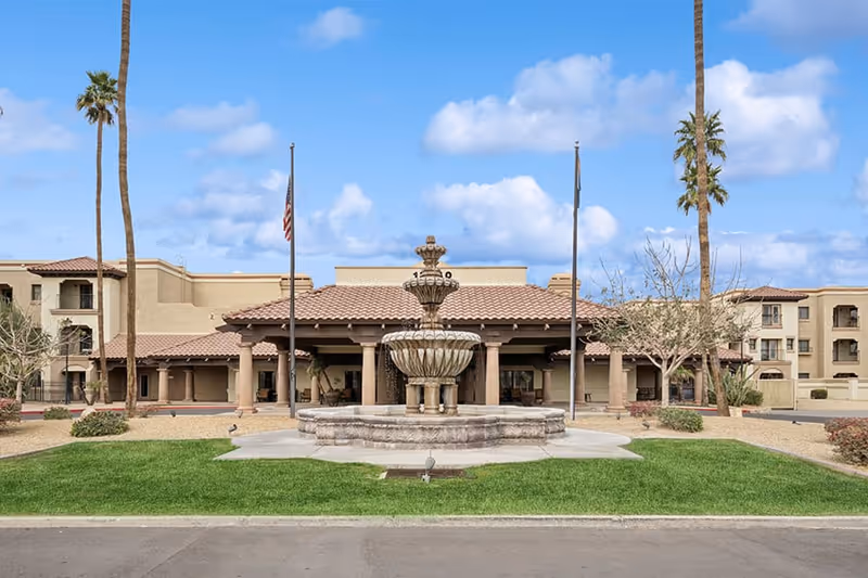 Front exterior view of The Forum at Desert Harbor senior living facility featuring a large stone fountain in the center, two flagpoles, palm trees, and a beige stucco building with a tiled roof under a partly cloudy blue sky.