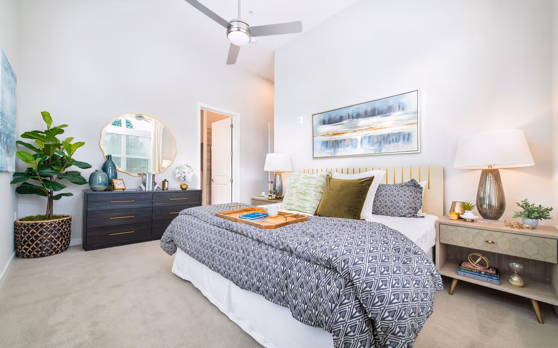 Bright and modern bedroom with a large bed covered in patterned bedding and multiple pillows. A wooden tray with a book and a cup sits on the bed. On the right side, there is a nightstand with a lamp, small plants, and decorative items. On the left side, a dark dresser with gold handles holds decorative vases, books, and a round mirror above it. A large potted plant is placed next to the dresser. The room has light-colored walls, carpeted floor, and a ceiling fan with light.