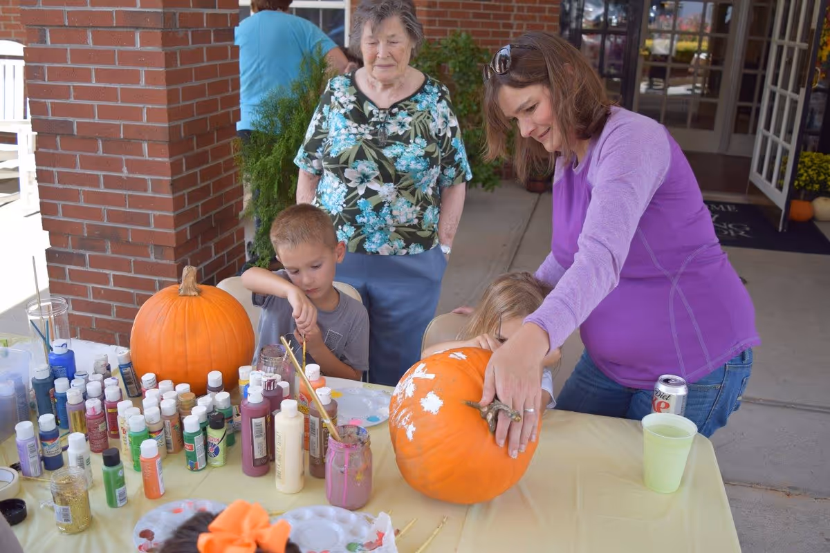 A woman in a purple long-sleeve shirt helps a young child paint a large pumpkin on a table covered with various bottles of paint and brushes. A young boy is also painting a pumpkin nearby, while an elderly woman in a floral shirt watches them. The scene takes place outside a brick building with an open door in the background.