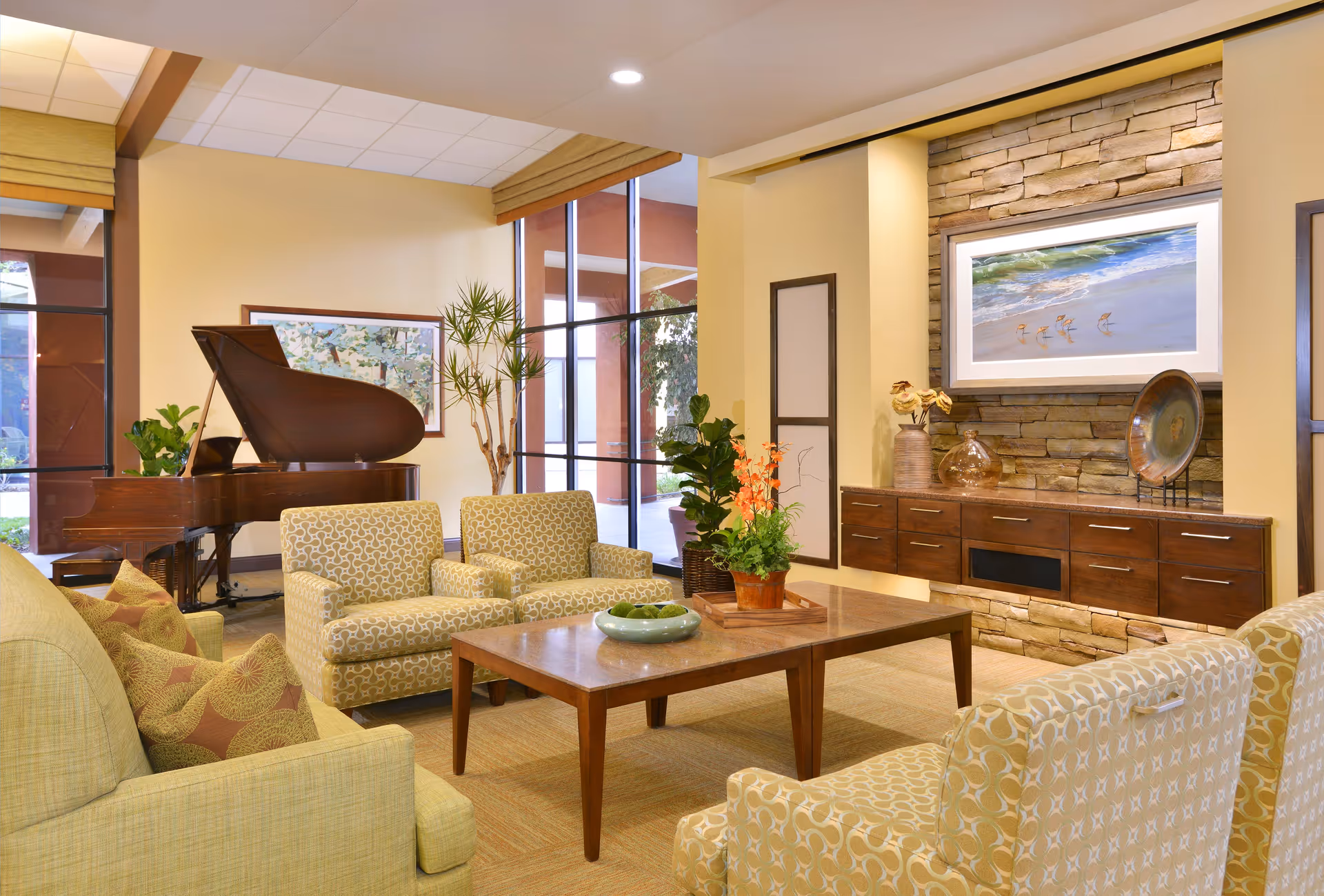 A cozy living room area in a retirement community featuring a grand piano, patterned armchairs, a wooden coffee table with a decorative bowl and plants, a stone accent wall with a mounted flat-screen TV, and large windows letting in natural light.
