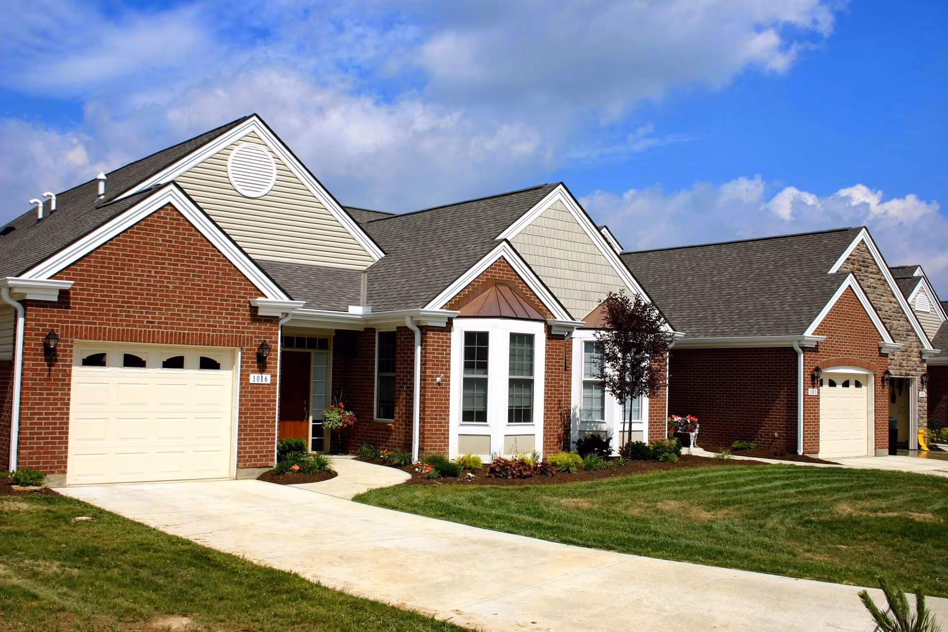 Exterior view of a single-story brick residential building with attached garages, manicured lawns, and a clear blue sky with some clouds.