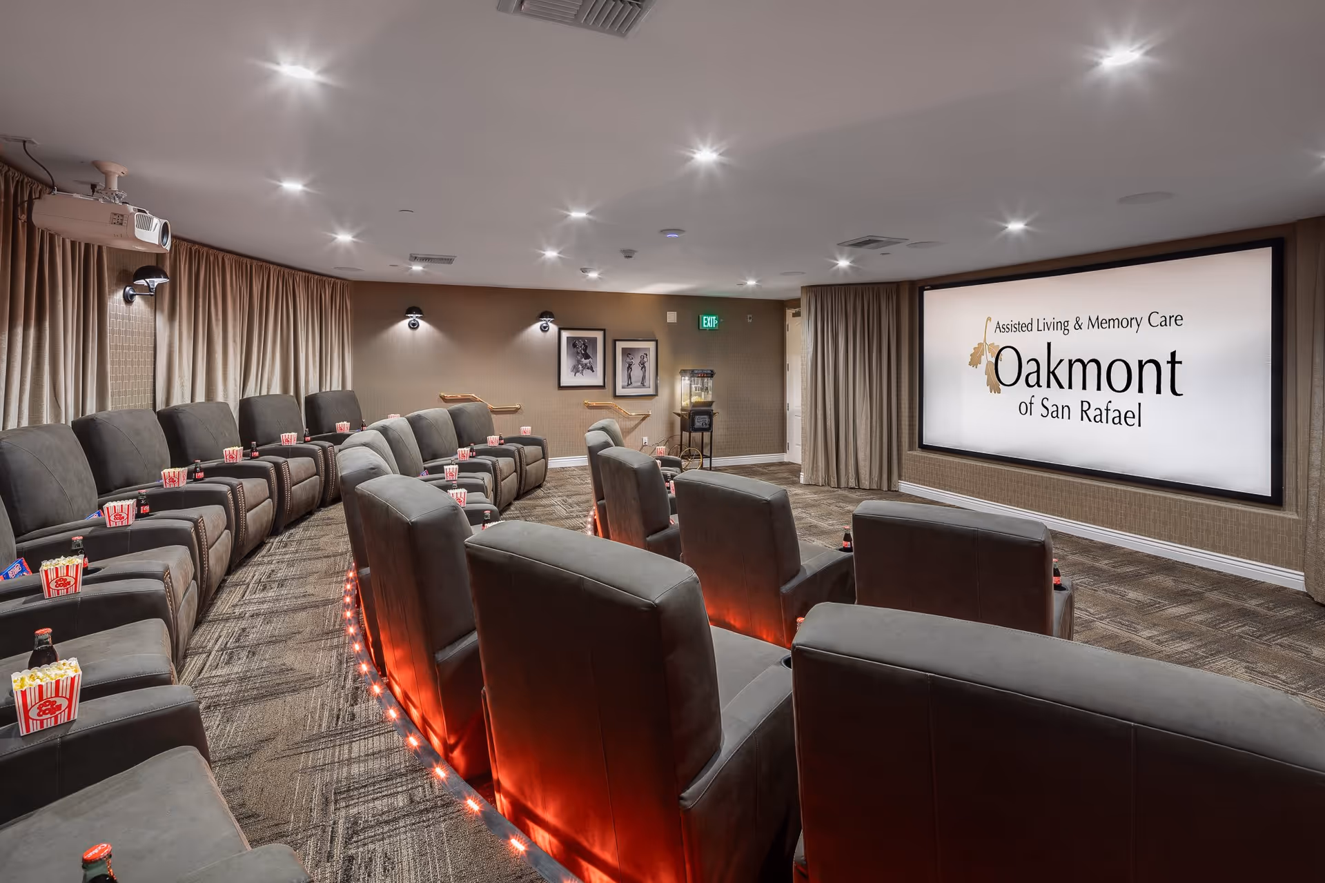 A cozy, modern theater room with two rows of gray reclining seats arranged in a curved layout facing a large screen displaying the text 'Assisted Living & Memory Care Oakmont of San Rafael.' Each seat has a popcorn container and a bottle of soda. The room features carpeted floors, wall sconces, framed pictures, a popcorn machine in the corner, and ceiling-mounted projector.