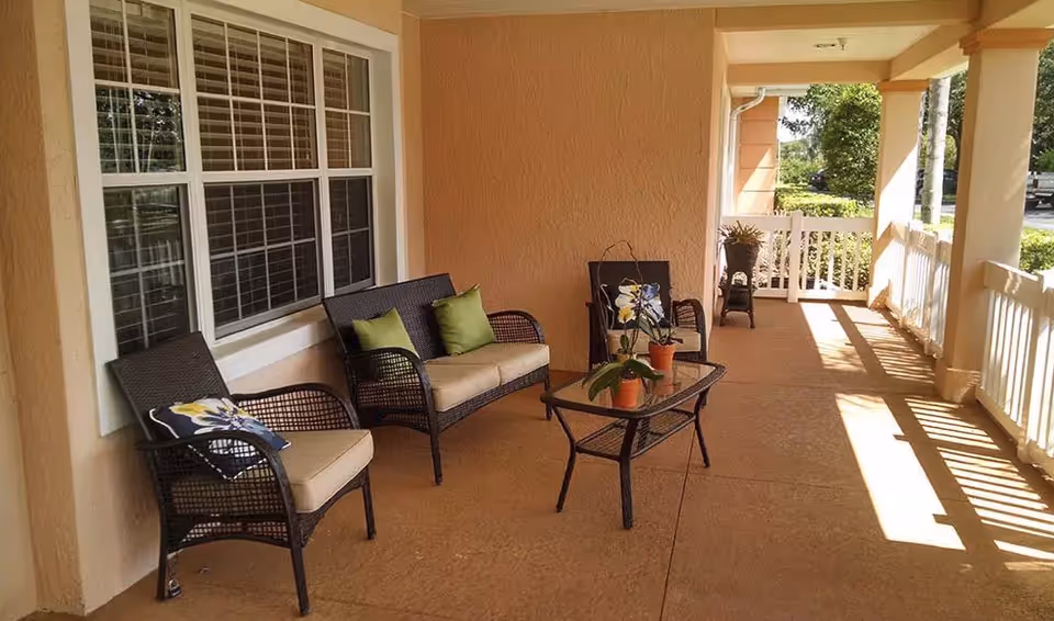 A covered outdoor porch area with a cushioned wicker loveseat, two cushioned wicker chairs, and a glass-top wicker coffee table with potted plants. The porch has beige walls, a large window with white trim, and white railing along the edge. Sunlight casts shadows on the floor and railing.