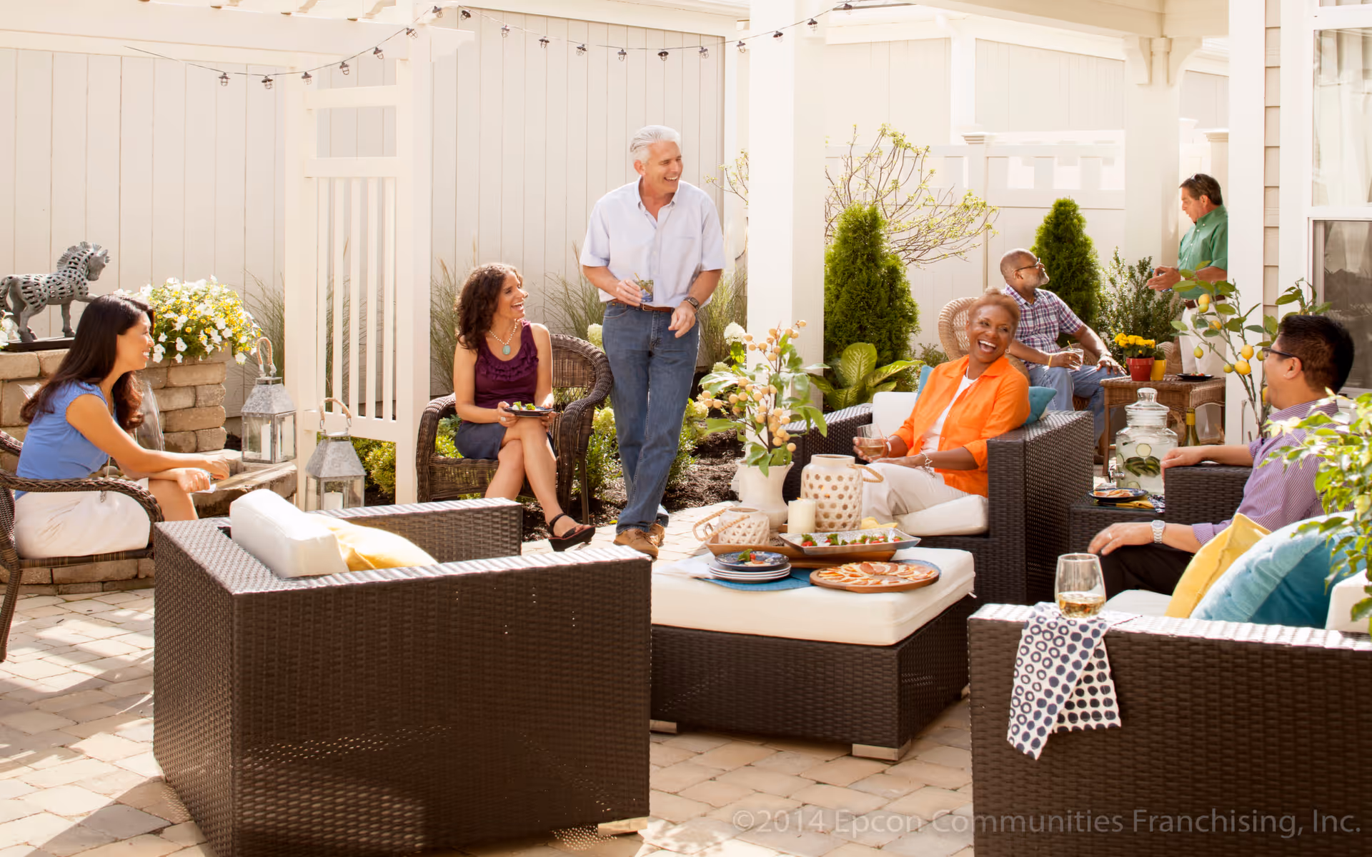 A group of six adults socializing and relaxing in a sunny outdoor patio area with wicker furniture, plants, and decorative items. They are smiling and enjoying drinks and snacks in a fenced garden space.