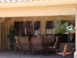 Covered outdoor patio area with cushioned chairs arranged around a small table, surrounded by windows and a hanging basket with flowers.