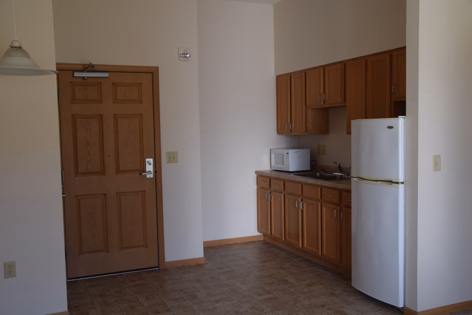 Small kitchenette with wooden cabinets, a white refrigerator and microwave beside a wooden entry door.