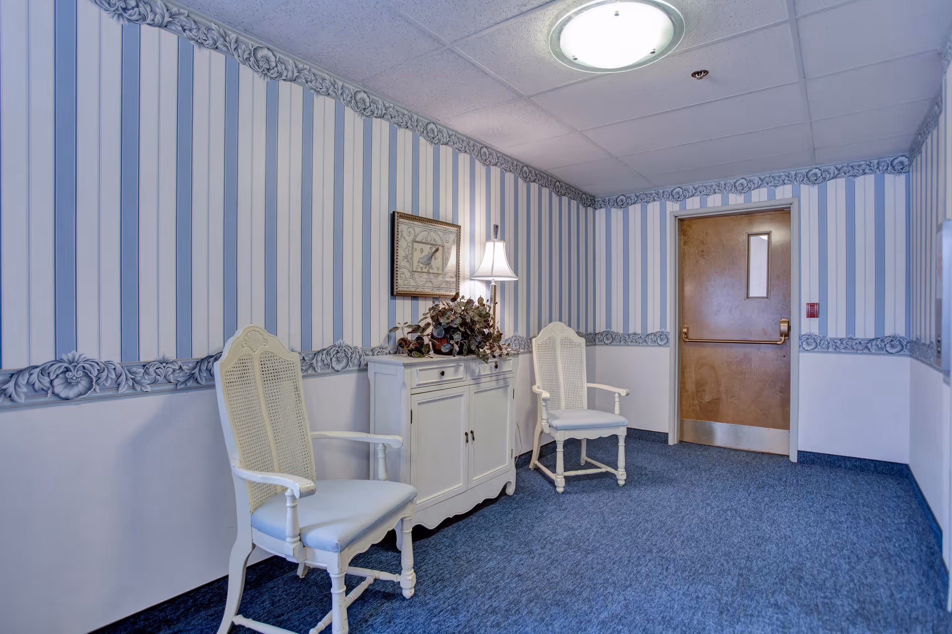 A small seating area in a hallway with two white wooden chairs with armrests, a white cabinet with a lamp and a plant on top, blue carpet, striped wallpaper with floral border, and a wooden door with a small window.