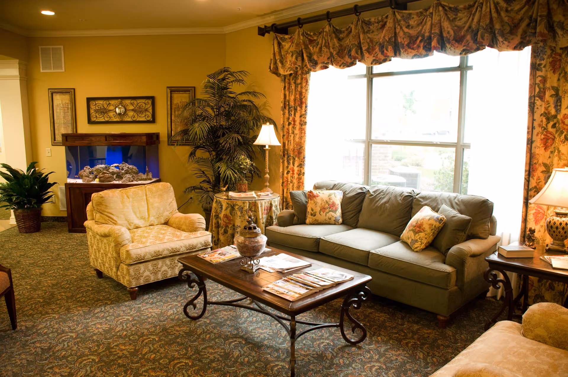 A cozy living room in a senior living facility with a green sofa adorned with floral pillows, a beige armchair, a wooden coffee table with magazines, a side table with a lamp and books, a large window with floral curtains, a potted plant, and an aquarium against a yellow wall.