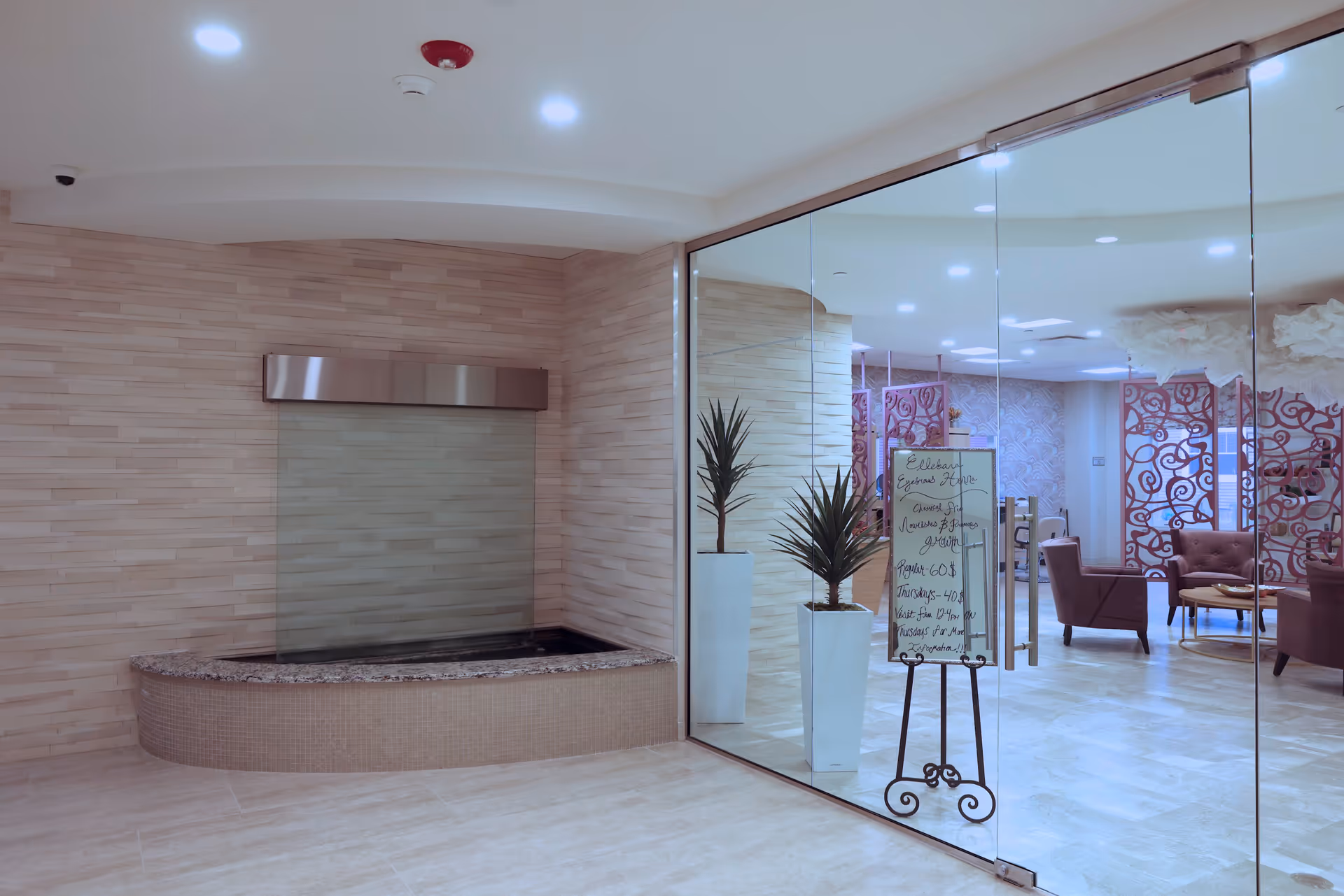 Glass-walled modern interior lobby with an indoor wall fountain, potted plants and seating visible through the doors.