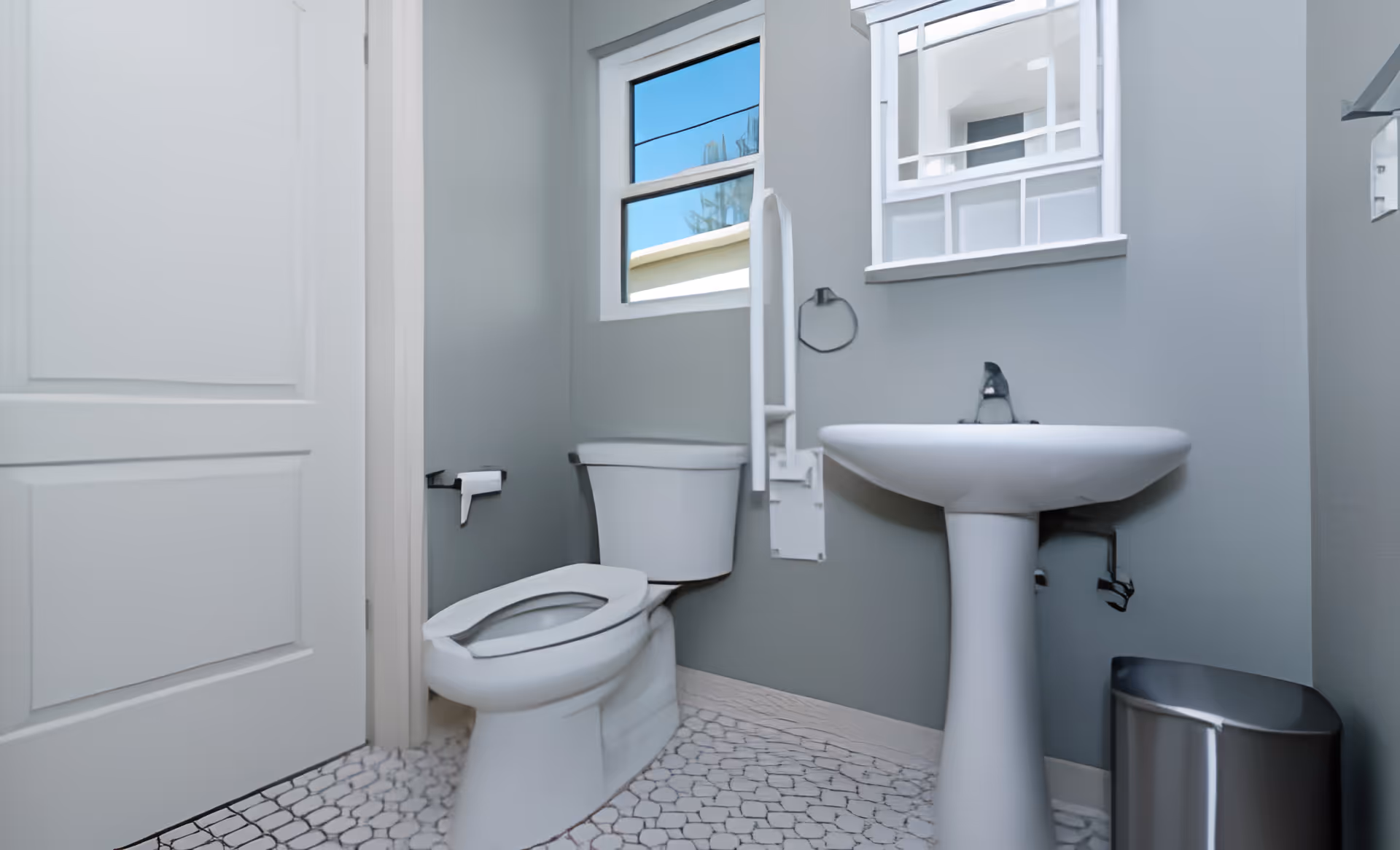 Small accessible bathroom with a toilet, pedestal sink, wall-mounted grab bar, window and mirror, and a trash can.