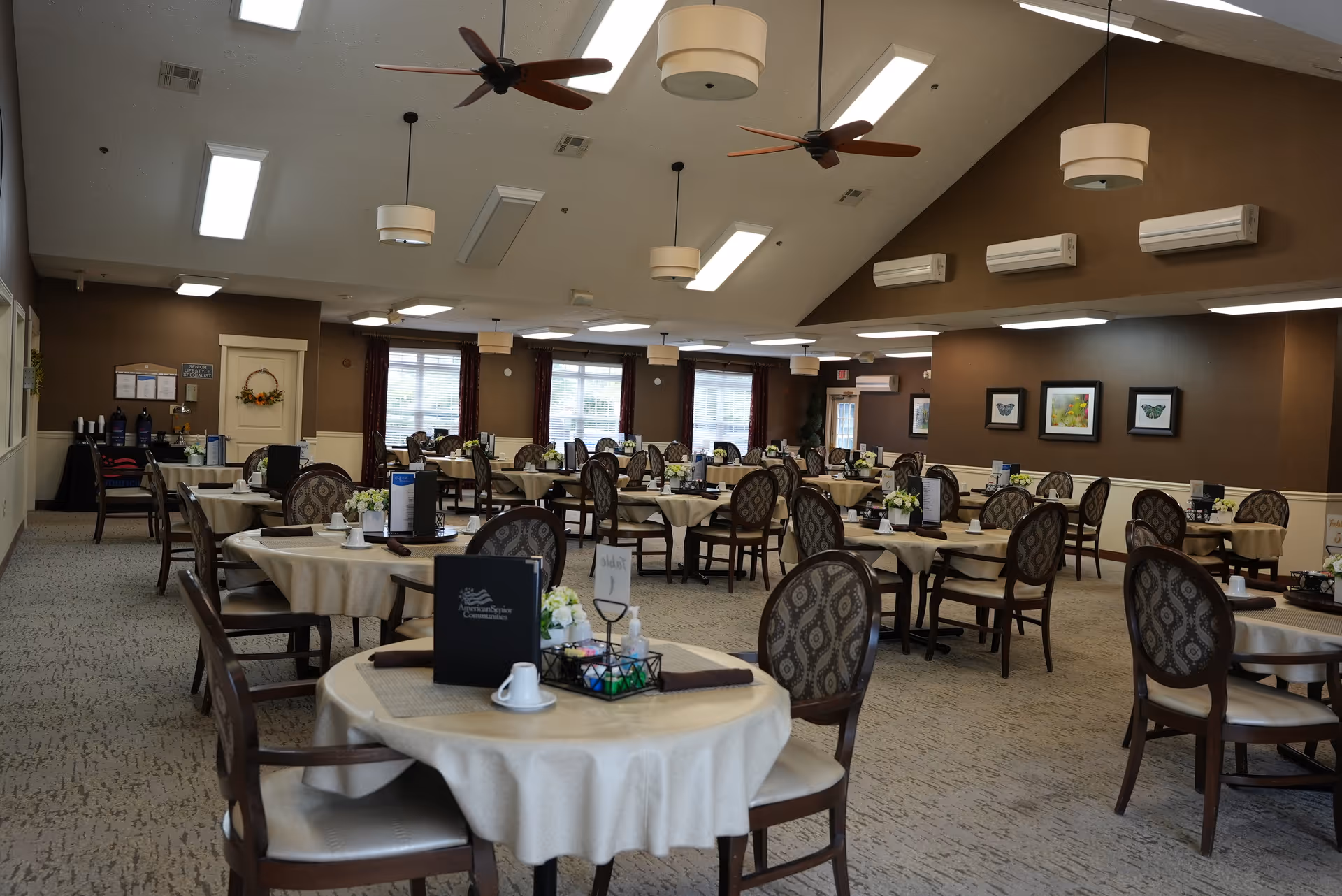 A spacious dining room with multiple round tables covered with beige tablecloths. Each table has chairs with patterned upholstery, small flower arrangements, menus, and place settings. The room has large windows with curtains, ceiling fans, and pendant lights hanging from a high ceiling. The walls are decorated with framed artwork and the room has a warm, inviting atmosphere.