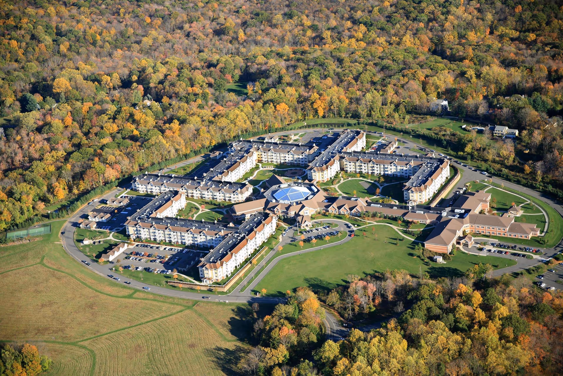 Aerial view of Meadow Ridge senior living facility surrounded by trees with autumn foliage and open green spaces. The large building complex has multiple wings, parking lots, and a central circular area with a glass roof.