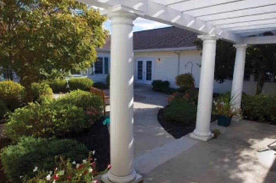 Outdoor garden area with white columns supporting a pergola, surrounded by green bushes, trees, and flowering plants. A building with white siding and a brown roof is visible in the background.