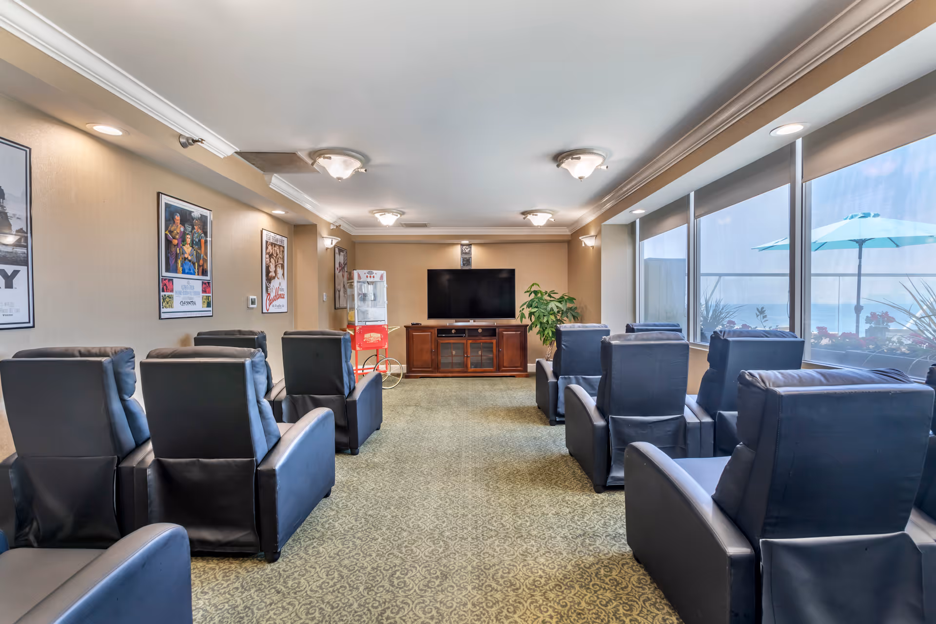 A cozy common area with multiple black leather recliners arranged in two rows facing a flat-screen TV on a wooden cabinet. The room has beige walls adorned with framed movie posters, a popcorn machine in the corner, and large windows on the right side letting in natural light with a view of an outdoor patio umbrella and plants.