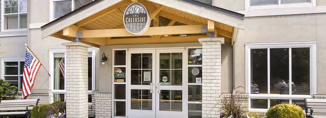Front entrance of The Creekside building with double glass doors, a circular 'The Creekside' sign above the doorway, and an American flag nearby.
