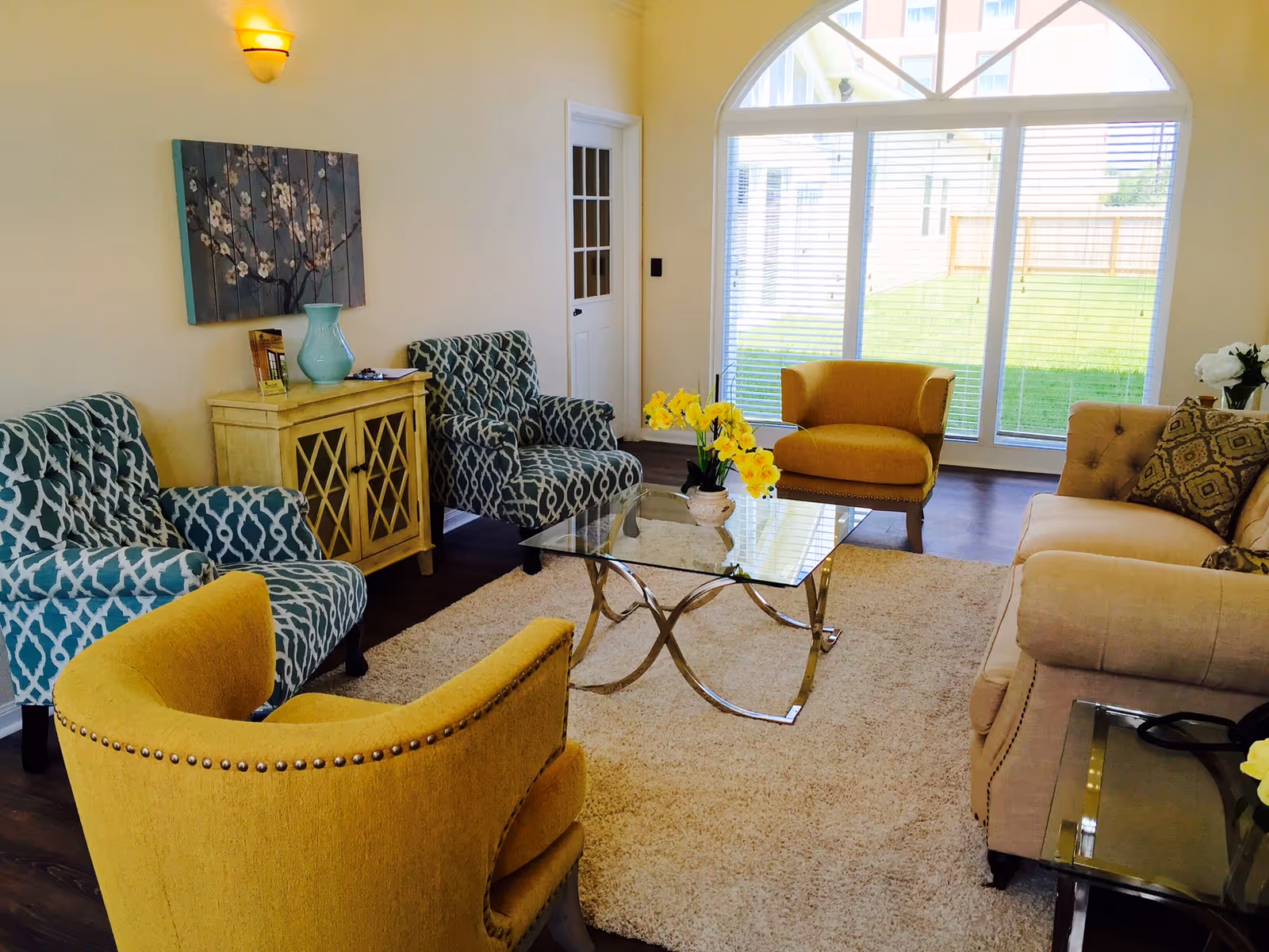 A bright living room with large arched windows letting in natural light. The room features a beige sofa with patterned cushions, two yellow armchairs, and two blue patterned armchairs arranged around a glass coffee table with a vase of yellow flowers. A light-colored rug covers the dark wooden floor, and a small wooden cabinet with a blue vase and decorative items is against the wall beneath a floral painting.