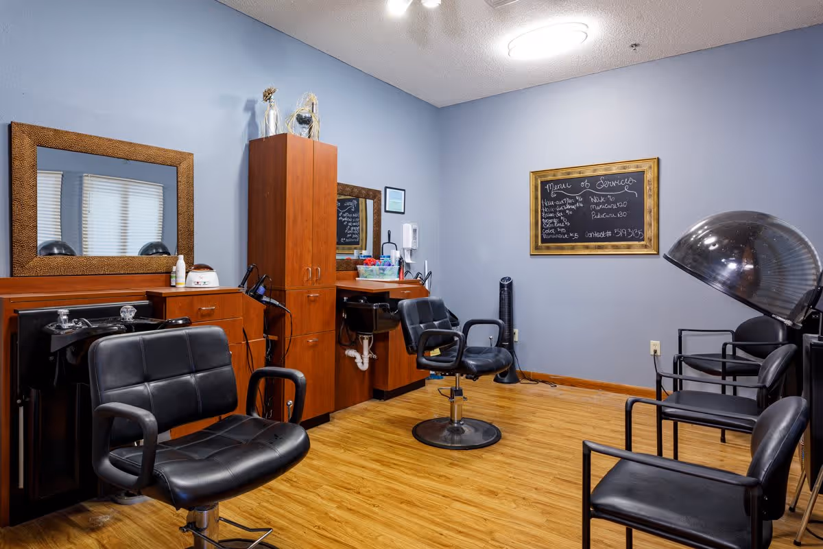 Interior of a hair salon room with light blue walls and wooden flooring. The room contains black salon chairs, a hair washing station with sinks, a tall wooden cabinet, a wall-mounted mirror with a decorative frame, and a blackboard menu listing services and prices. There is also a hair dryer hood and additional seating along the right wall.