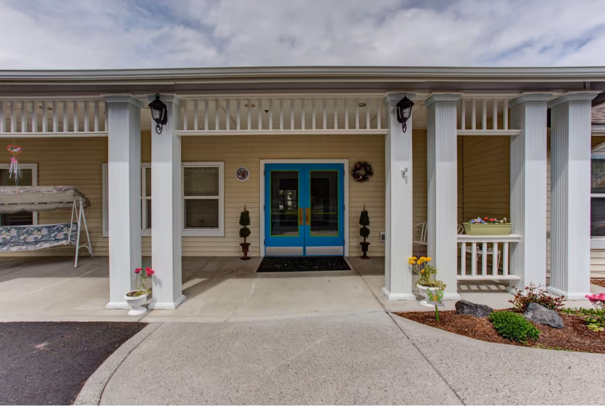 Covered front entrance of a memory care building featuring blue double doors framed by white columns, potted plants, and a porch swing.