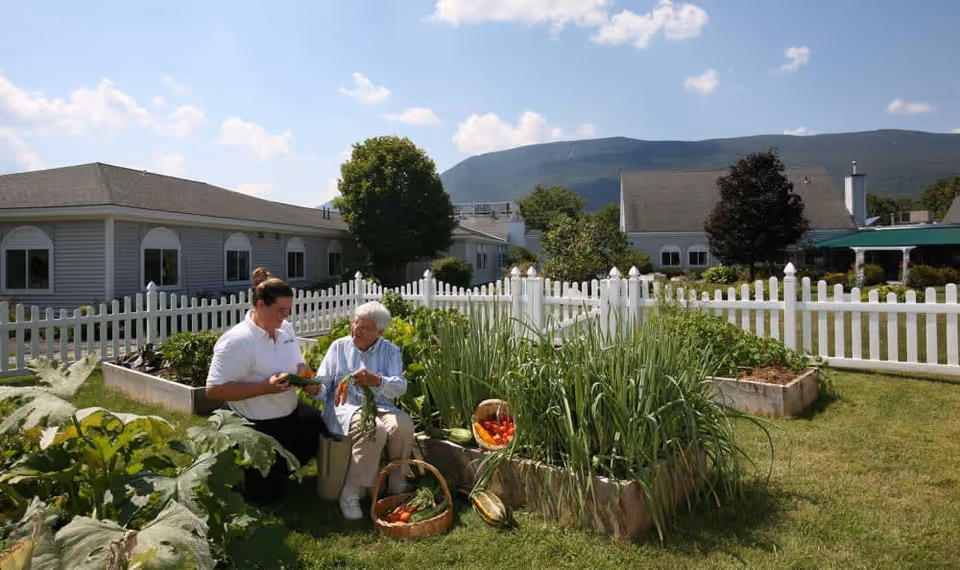 An elderly woman and a caregiver are sitting outdoors in a garden area with raised planting beds filled with vegetables and plants. They are holding freshly picked vegetables and appear to be engaged in a pleasant conversation. The garden is enclosed by a white picket fence, with residential buildings and mountains visible in the background under a partly cloudy sky.